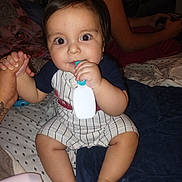 Roberto is registered to the contest to win money with this photo: baby, child, infant, person, hand, bed, blanket, clothing, toy, chewing, sitting, indoor, adult, game_controller, holding, skin, face, eyes, foot, comfort