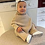 baby, child, infant, smile, headband, bow, knitwear, beige, white_shoes, ottoman, indoor, kitchen, furniture, happy, cute, seated, portrait, cozy, apparel, person