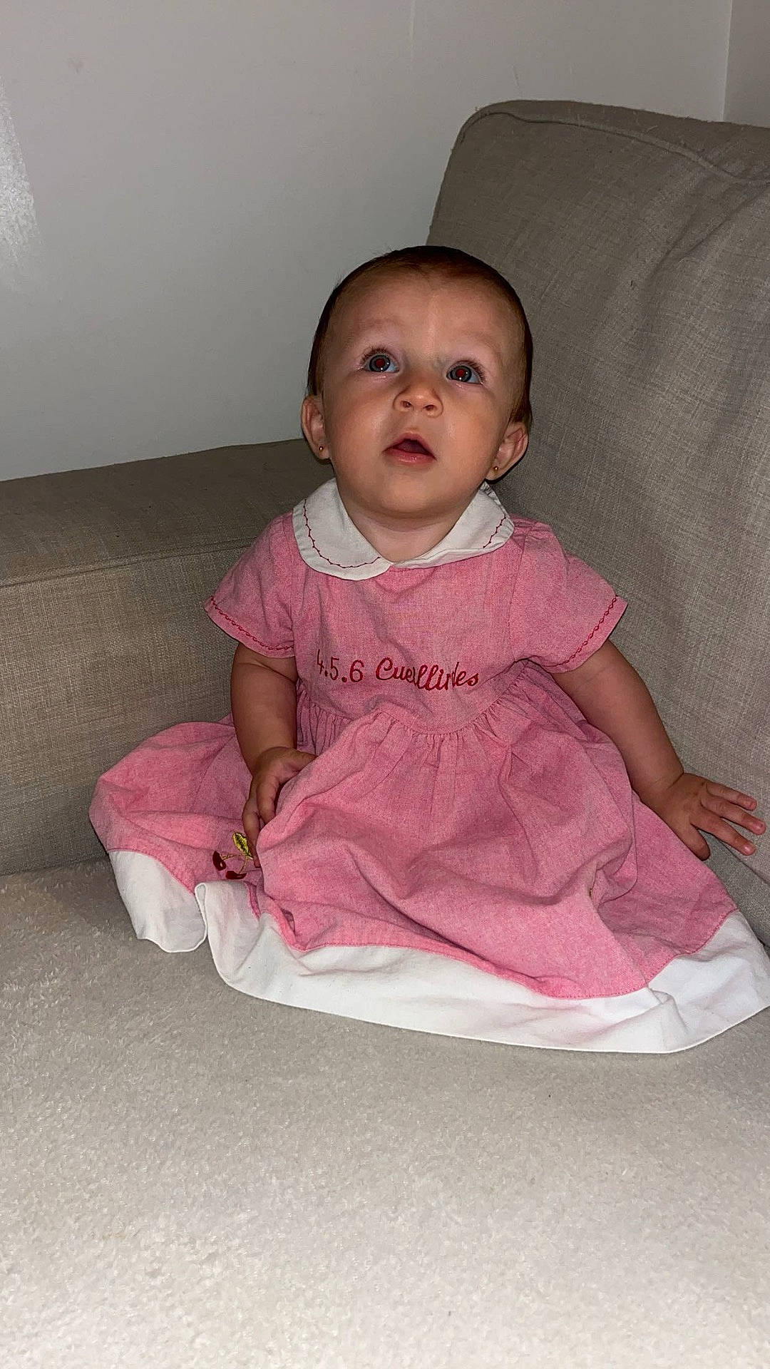child, collar, couch, curious, dress, earrings, expression, fabric, face, furniture, home, indoor, looking_up, person, pink, short_hair, sitting, toddler, wall, young_child