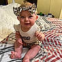 baby, child, infant, smiling, headband, bow, bed, bedding, patterned, clothing, sitting, cute, indoors, person, happy, skin, foot, hand, room, text_on_clothing