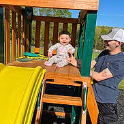Izzy joined the competition — help win amazing prizes! baby, child, playground, slide, wooden_structure, outdoor, adult, smiling, holding_hand, sunny, greenery, park, casual_clothing, hat, tattoos, shorts, daylight, happy, play, support