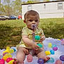 Izzy joined the competition — help win amazing prizes! baby, pacifier, easter_eggs, blanket, grass, outdoor, child, toy, yellow_clothing, sitting, feet, spring, daylight, mobile_home, cars, greenery, playtime, cute, colorful, nature