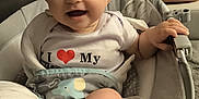 Inaya a rejoint le concours — aidez-le/la à gagner de superbes lots ! baby, infant, smiling, child, white_shirt, heart_graphic, baby_seat, cushion, indoor, carpet, happy, cute, chubby_legs, hand, foot, face, person, young_child, comfortable, seat