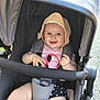 baby, child, stroller, smile, hat, bonnet, sunglasses, pink_shirt, black_shorts, daisy_pattern, seatbelt, outdoor, sunlight, happy, cute, infant, person, summer, playful, hands