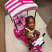 Yomi is registered to the contest to win money with this photo: child, girl, toy_scooter, pink, indoor, smiling, braided_hair, beads, floor_tile, playful, canopy, toy, vehicle, happy, person, cute, colorful, young, small, seated