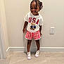 Yomi is registered to the contest to win money with this photo: toddler, child, smile, braids, beads, tshirt, shorts, shoes, floor, wall, indoor, power_outlet, standing, happy, casual_clothing, person, portrait, cute, fashion, accessory