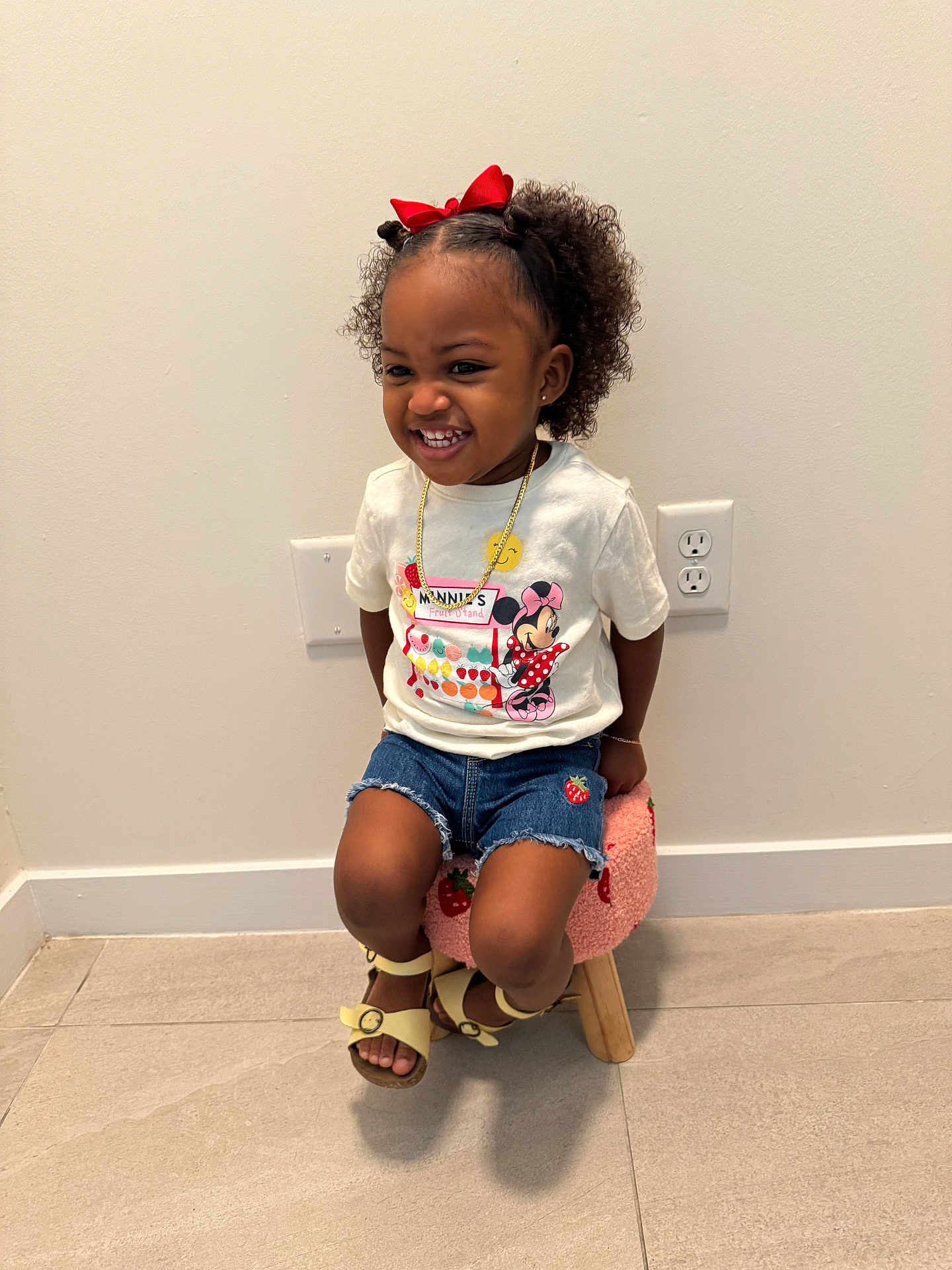 Yomi is registered to the contest to win money with this photo: child, girl, smiling, curly_hair, red_bow, minnie_mouse, tshirt, denim_shorts, strawberry_patch, stool, indoor, floor_tiles, wall_outlet, sandals, happy, cute, sitting, jewelry, gold_chain, playful