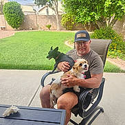 Roxy And Stella is registered to the contest to win money with this photo: man, dog, black_dog, brown_white_dog, chair, patio, backyard, grass, tree, shrub, fence, table, cap, smile, outdoor, sunlight, relaxation, pet, leisure, summer