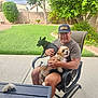 man, dog, black_dog, brown_white_dog, chair, patio, backyard, grass, tree, shrub, fence, table, cap, smile, outdoor, sunlight, relaxation, pet, leisure, summer