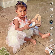 Gursahib is registered to the contest to win money with this photo: child, toddler, girl, dress, pink, white, sitting, indoor, tile_floor, toy, wooden_toy, sandals, hair_bows, portrait, looking_at_camera, home, dining_area, rug, social_media_screenshot, smartphone_interface