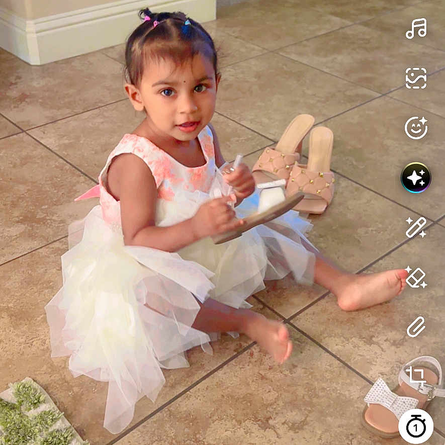 Gursahib is registered to the contest to win money with this photo: child, toddler, girl, dress, pink, white, sitting, indoor, tile_floor, toy, wooden_toy, sandals, hair_bows, portrait, looking_at_camera, home, dining_area, rug, social_media_screenshot, smartphone_interface