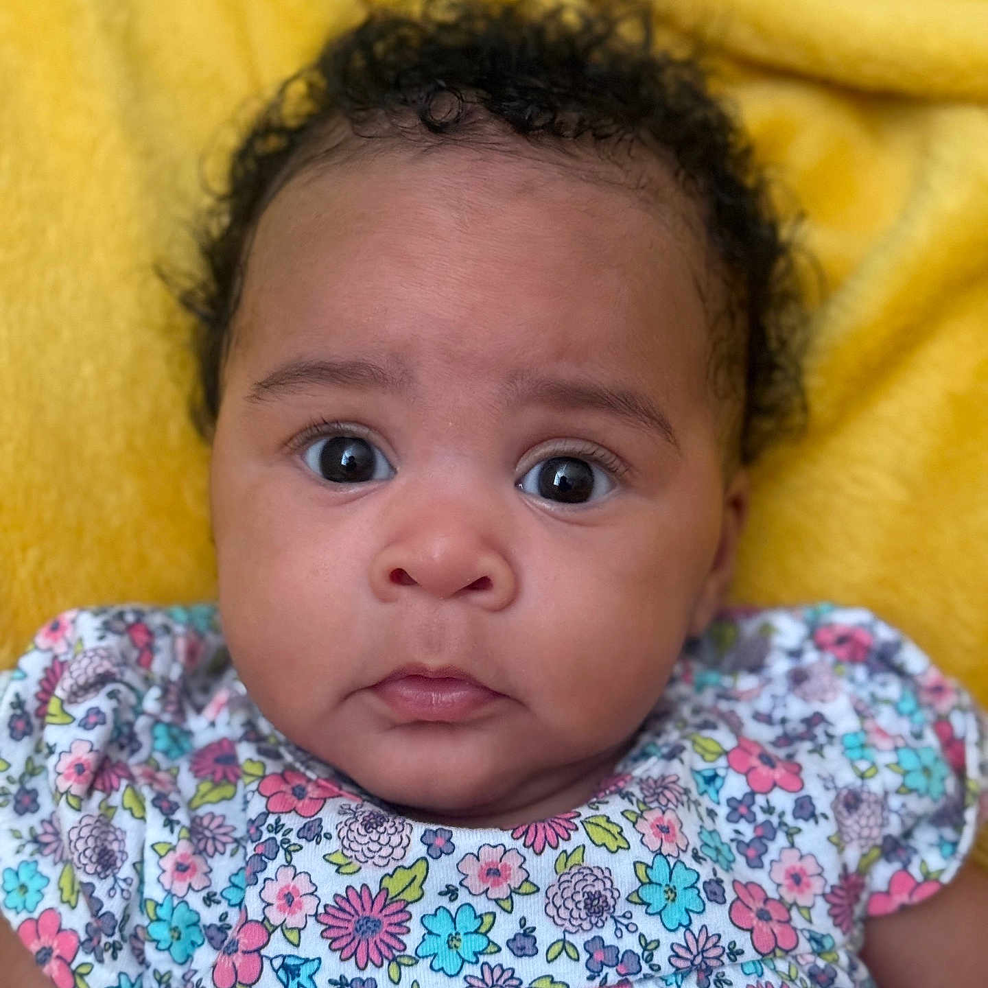 Moon is registered to the contest to win money with this photo: adorable, baby, child, closeup, colorful, curly_hair, cute, expression, face, floral_clothing, indoors, infant, person, portrait, sitting, skin, soft_texture, wide_eyes, yellow_blanket, young