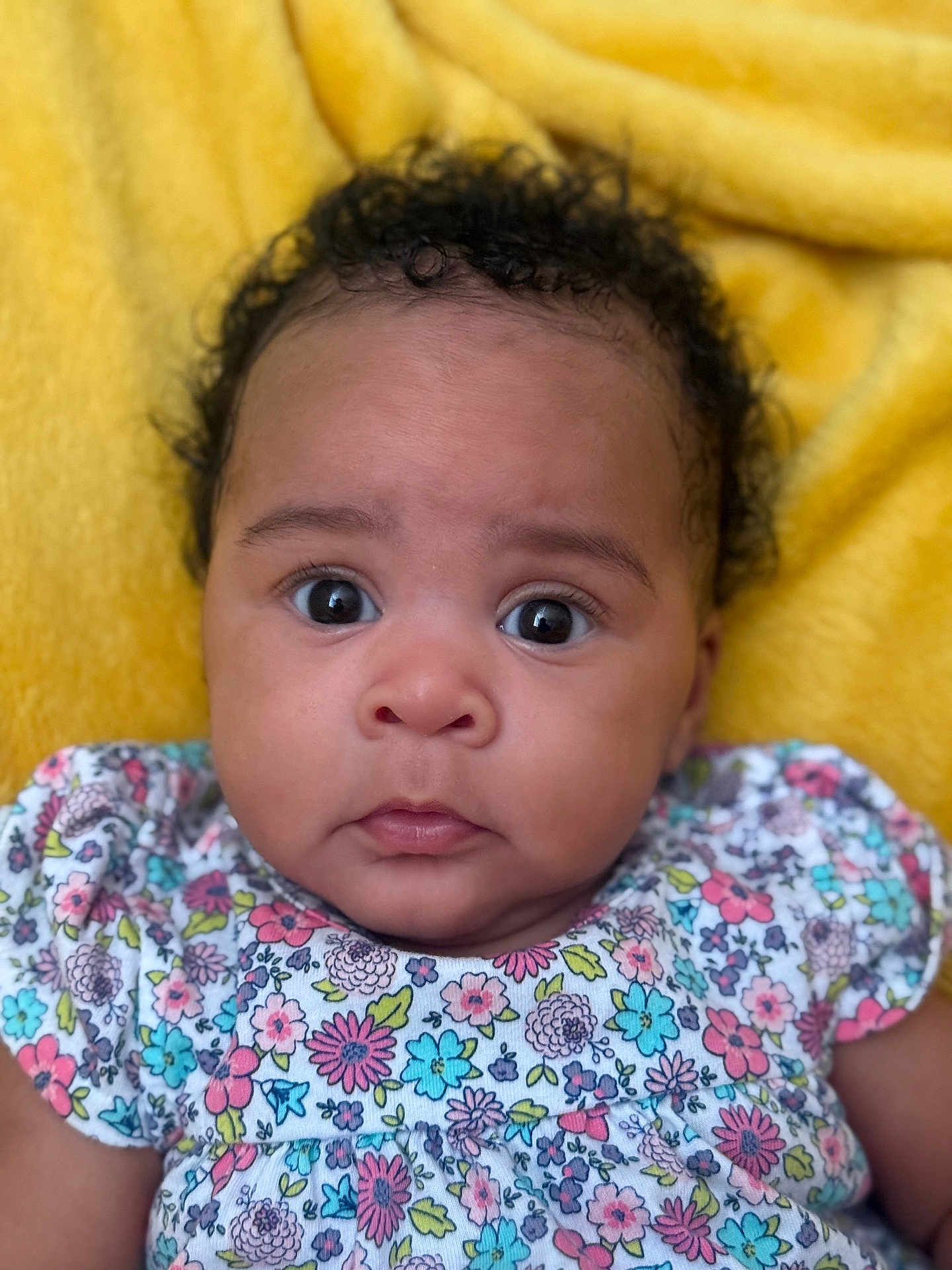 Moon is registered to the contest to win money with this photo: baby, infant, face, curly_hair, wide_eyes, floral_clothing, colorful, yellow_blanket, portrait, cute, child, young, closeup, expression, skin, person, sitting, indoors, soft_texture, adorable