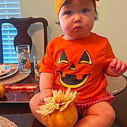 Katherine is registered to the contest to win money with this photo: baby, child, orange_clothing, headband, pumpkin, table, flower, sitting, indoor, dining_room, chair, glasses, plate, placemat, cute, festive, holiday, face, person, foot