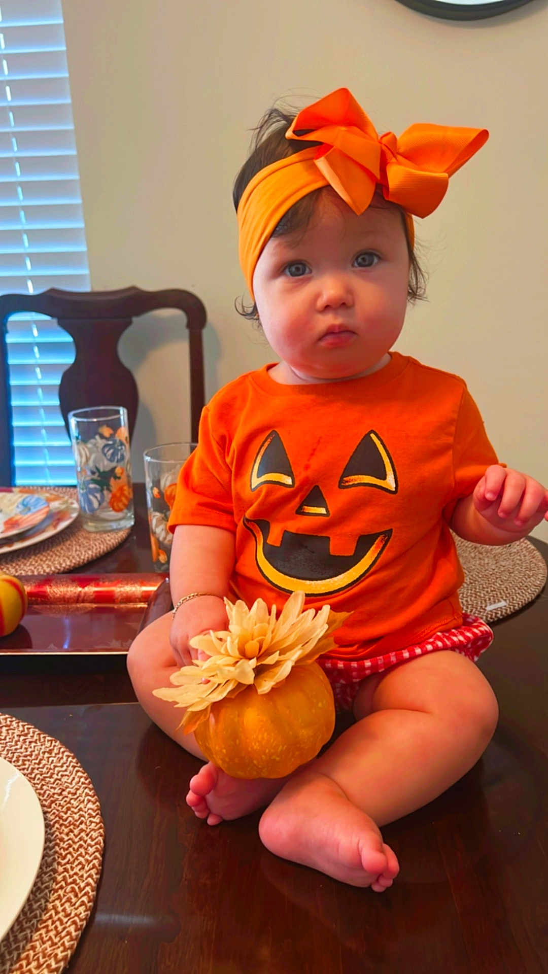 Katherine is registered to the contest to win money with this photo: baby, child, orange_clothing, headband, pumpkin, table, flower, sitting, indoor, dining_room, chair, glasses, plate, placemat, cute, festive, holiday, face, person, foot