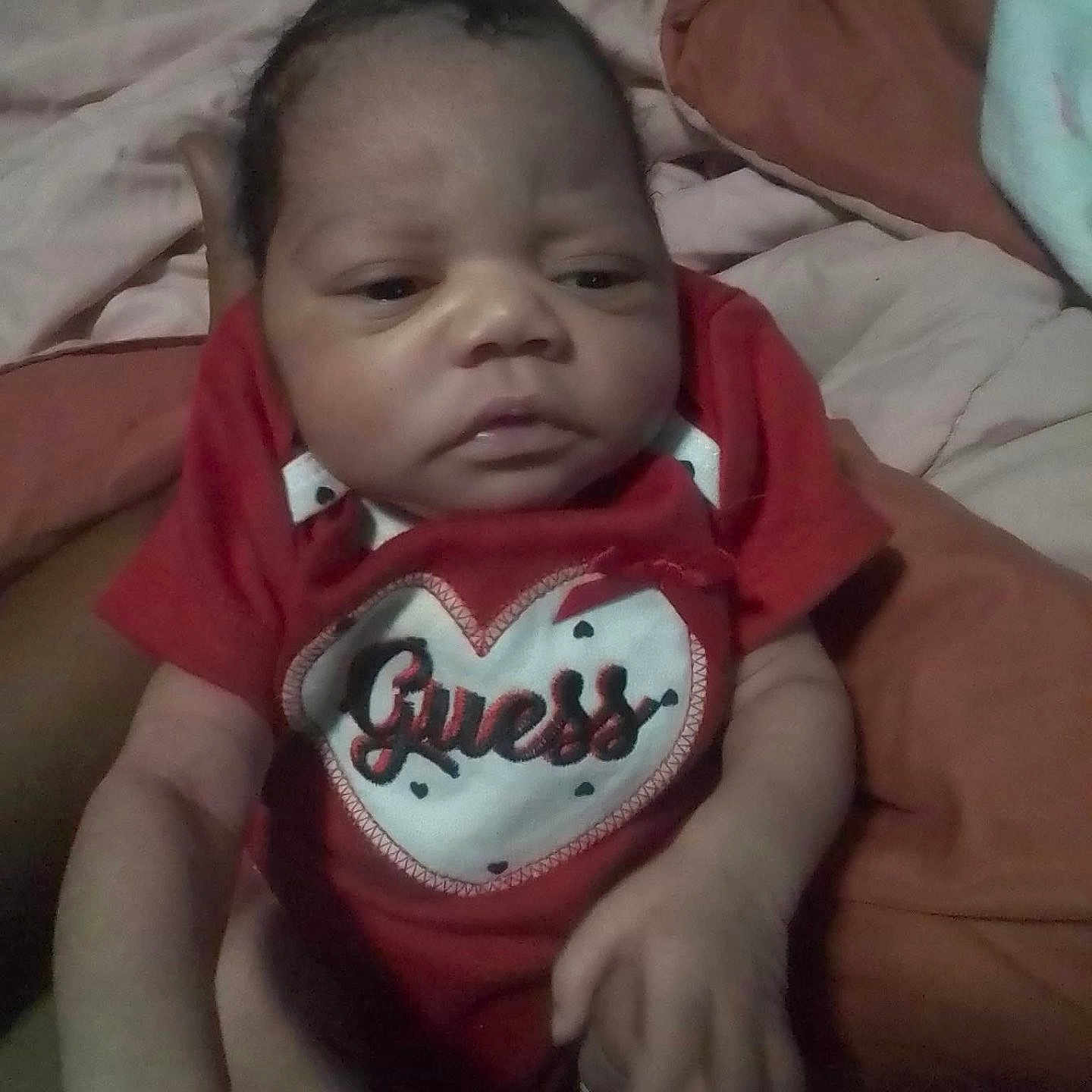 Carlynn is registered to the contest to win money with this photo: baby, infant, red_onesie, clothing, heart_logo, guess_brand, person, hand, blanket, cushion, bed, indoor, portrait, newborn, cute, skin, face, expression, holding, soft