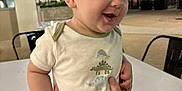 Jackson is registered to the contest to win money with this photo: baby, child, infant, onesie, dinosaur, hands, tattoo, table, outdoor, night, smile, happy, person, restaurant, light, face, clothing, skin, holding, background