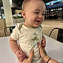 baby, child, infant, onesie, dinosaur, hands, tattoo, table, outdoor, night, smile, happy, person, restaurant, light, face, clothing, skin, holding, background