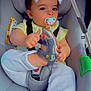 baby, child, car_seat, pacifier, sandal, footwear, white_pants, yellow_shirt, seatbelt, car_accessory, cute, infant, indoors, playful, face_filter, cat_ears, small_child, holding, sitting, straps