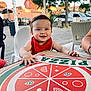 arm, baby, building, car, casual, child, daylight, face, hand, happy, high_chair, outdoor, person, pizza_box, red_bib, seat, sidewalk, smiling, table, tree