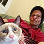 Runny Babbit is registered to the contest to win money with this photo: cat, person, glasses, beanie, couch, indoor, whiskers, blue_eyes, face, hand, red_shirt, relaxed, furniture, living_room, portrait, pet, close_up, animal, casual, home