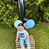 baby, boy, child, balloon, number_one, outdoor, grass, wooden_crate, cap, shirt, blue_balloon, white_balloon, smiling, barefoot, greenery, celebration, happy, casual_clothing, portrait, daylight