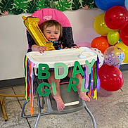 Addison is registered to the contest to win money with this photo: baby, high_chair, balloon, birthday, celebration, colorful, streamers, party, toddler, indoor, decorations, floor, smiling, person, foot, leaf_pattern, wall, chair, pink, number_one