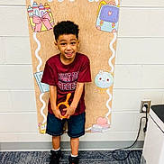 Rhyatt is registered to the contest to win money with this photo: child, boy, school, classroom, pre_k, smiling, standing, portrait, indoor, colorful, backpack, school_supplies, toy, orange_toy, red_shirt, shorts, sneakers, carpet, wall, classroom_decoration