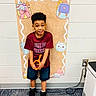child, boy, school, classroom, pre_k, smiling, standing, portrait, indoor, colorful, backpack, school_supplies, toy, orange_toy, red_shirt, shorts, sneakers, carpet, wall, classroom_decoration