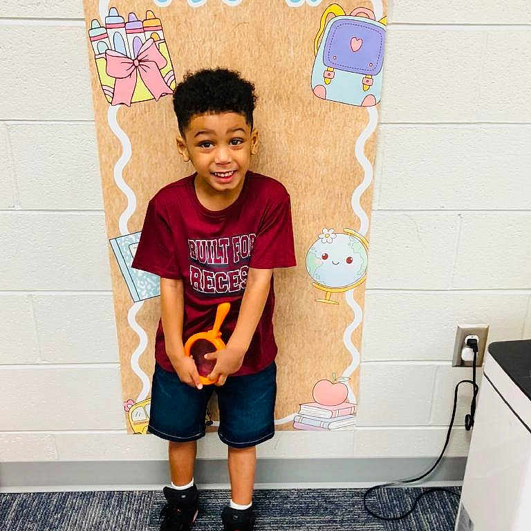 Rhyatt is registered to the contest to win money with this photo: child, boy, school, classroom, pre_k, smiling, standing, portrait, indoor, colorful, backpack, school_supplies, toy, orange_toy, red_shirt, shorts, sneakers, carpet, wall, classroom_decoration