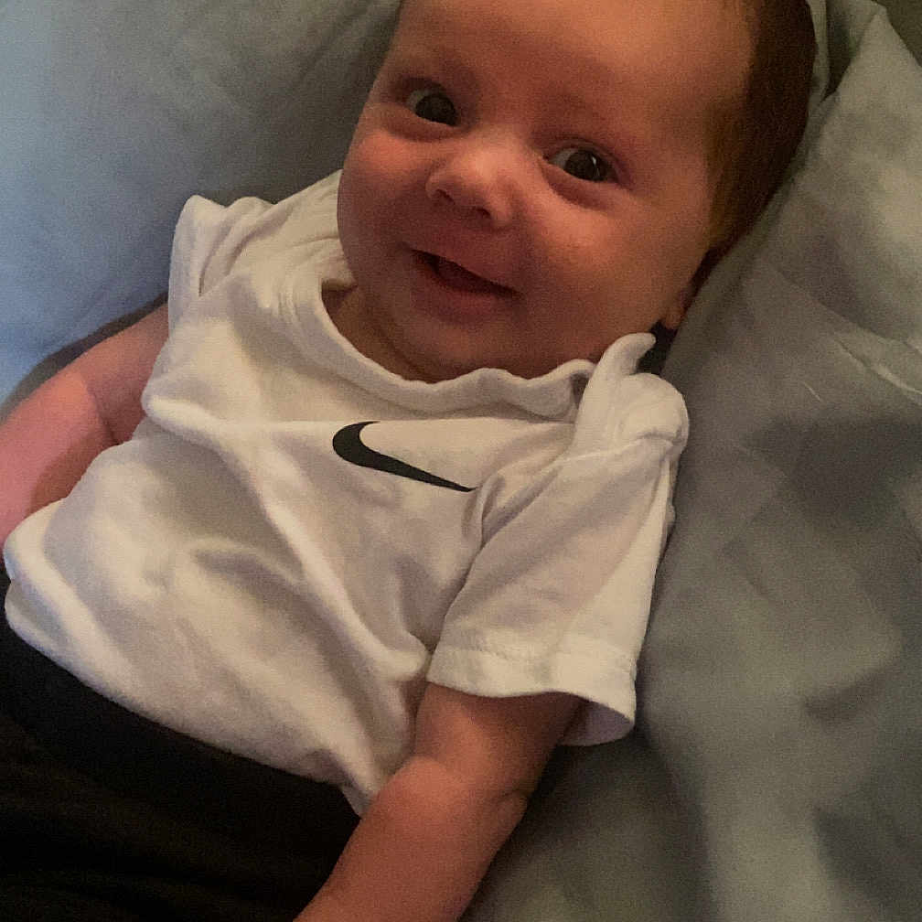 Marvin-John is registered to the contest to win money with this photo: baby, bed, blanket, casual_clothing, child, cute, face, hand, happy, indoors, infant, lying_down, nike_logo, pacifier, person, portrait, smiling, soft_light, white_tshirt, young