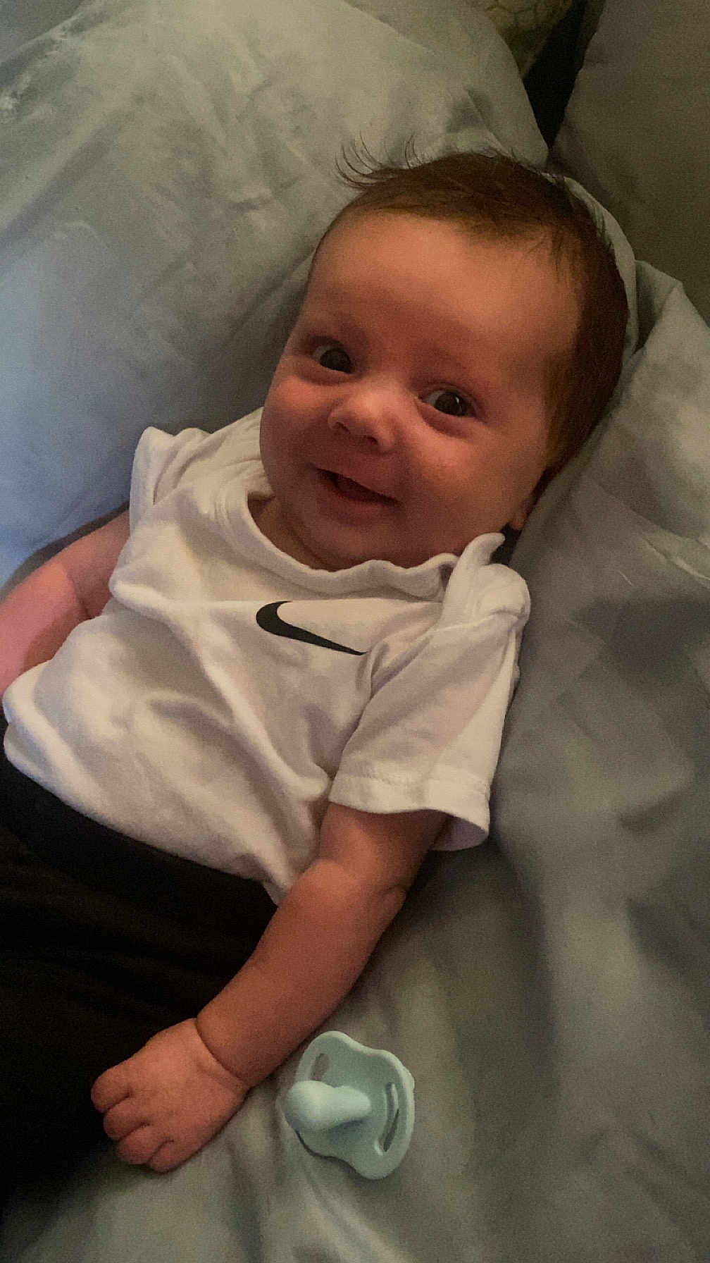 Marvin-John is registered to the contest to win money with this photo: baby, infant, smiling, white_tshirt, nike_logo, pacifier, bed, blanket, lying_down, cute, happy, child, young, person, indoors, soft_light, portrait, face, hand, casual_clothing