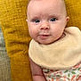 Eliza is registered to the contest to win money with this photo: baby, infant, child, face, eyes, smile, bib, clothing, floral, pillow, yellow, couch, skin, hand, portrait, indoors, cute, soft, young, person