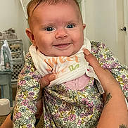 Eliza joined the competition — help win amazing prizes! baby, infant, smile, face, hands, tattoo, floral_clothing, onesie, bib, indoor, person, cute, child, closeup, expression, happy, holding, skin, portrait, young