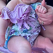 Eliza is registered to the contest to win money with this photo: baby, child, infant, floral_clothing, lavender, hat, blue_eyes, hand_holding, adult_hand, rings, nail_polish, inflatable_seat, pink, skin, face, legs, toes, person, closeup, outdoor