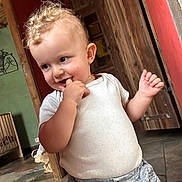 Anna participe au concours pour gagner de l'argent avec cette photo : toddler, child, indoor, curly_hair, white_shirt, gray_pants, wooden_door, floor_tiles, playful, smiling, person, cute, baby, standing, hand, face, expression, home, casual_clothing, young_child