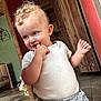 toddler, child, indoor, curly_hair, white_shirt, gray_pants, wooden_door, floor_tiles, playful, smiling, person, cute, baby, standing, hand, face, expression, home, casual_clothing, young_child