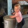 toddler, child, barefoot, curly_hair, pink_shirt, black_shorts, metal_container, handles, brick_wall, cage, water_bottle, cushion, floor, indoor, looking_back, curious, person, exploring, standing, young_child