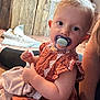baby, pacifier, blanket, child, dress, curly_hair, indoor, person, cute, infant, skin, face, hand, seated, looking, young_child, soft_toy, comfort, home, furniture
