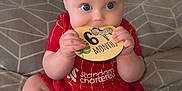 Willow is registered to the contest to win money with this photo: baby, infant, red_clothing, bed, blanket, plaque, wooden, sitting, chewing, cute, child, hands, feet, blue_eyes, patterned_bedspread, indoors, portrait, crawling_age, teething, smiling
