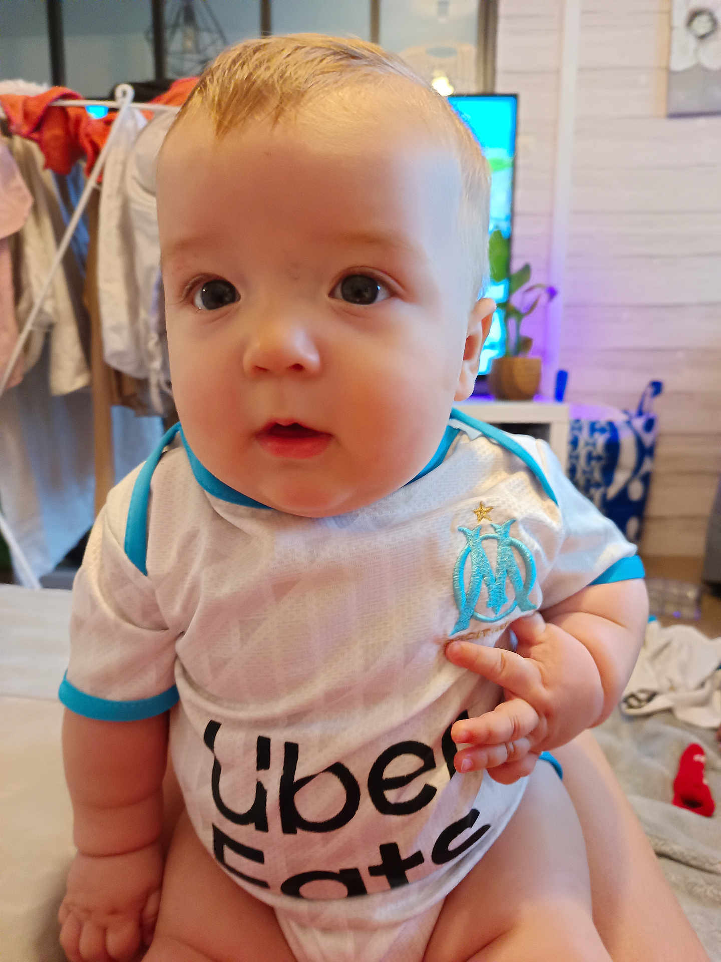 Sayan a rejoint le concours — aidez-le/la à gagner de superbes lots ! baby, child, infant, person, clothing, indoor, sports_jersey, uber_eats, curious, seated, skin, face, hand, furniture, laundry, background, home, blue, white, cute