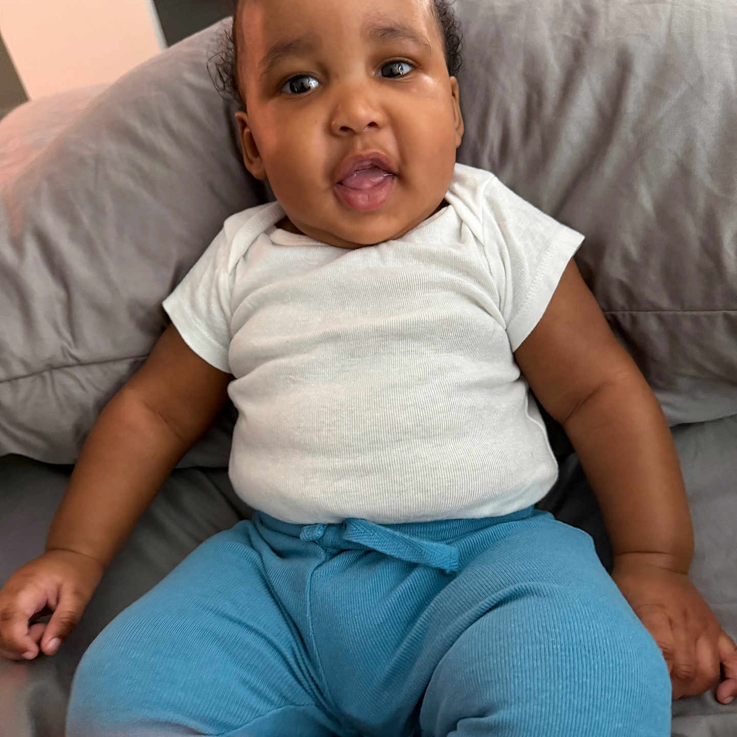 Micah is registered to the contest to win money with this photo: baby, child, infant, white_shirt, blue_pants, cushion, gray_cushion, sitting, indoors, curious_expression, cute, portrait, person, young_child, plump_arms, short_hair, skin, face, mouth_open, casual_clothing
