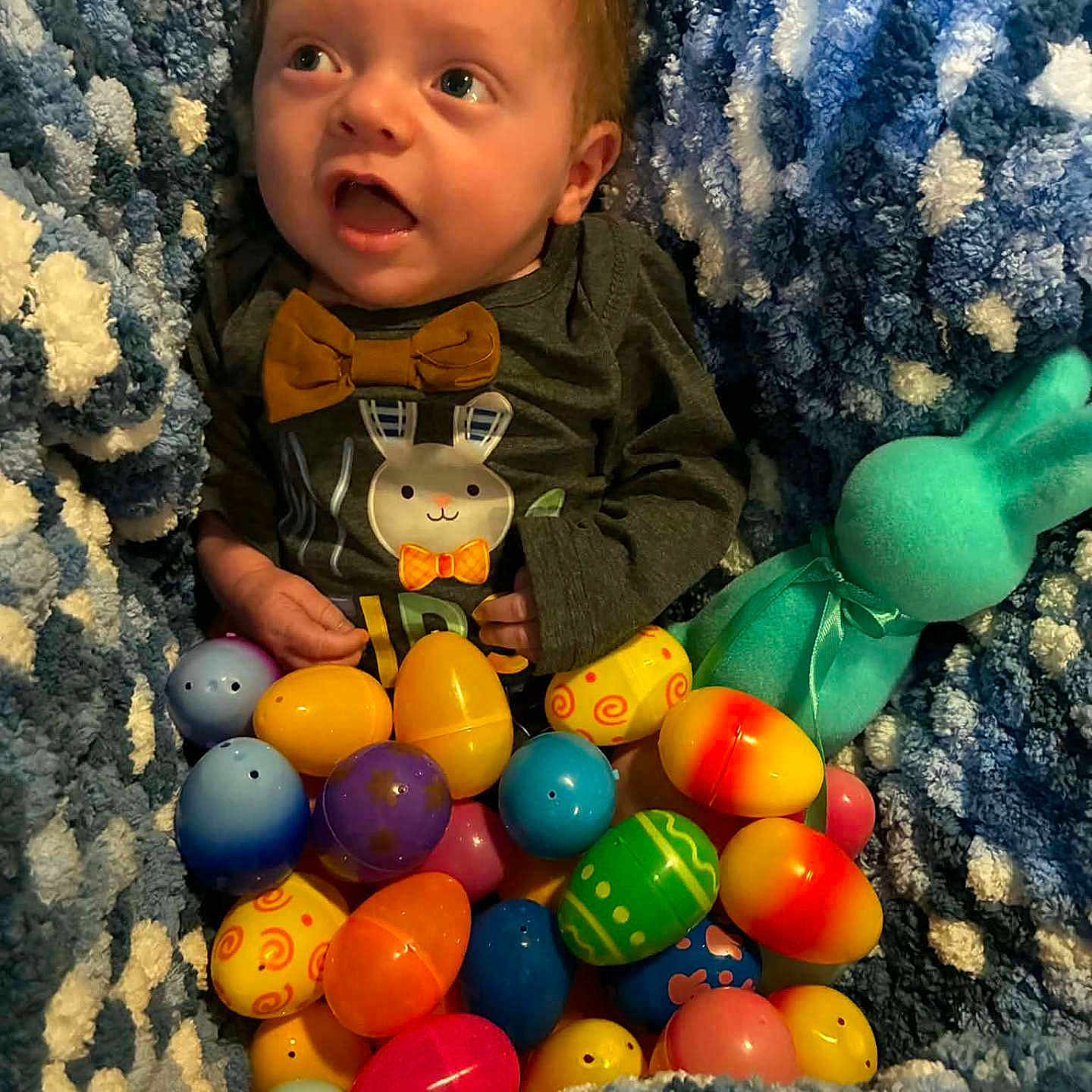 Roscowe is registered to the contest to win money with this photo: baby, child, easter_eggs, plush_toy, blanket, bunny, colorful, soft_texture, infant, cute, portrait, indoors, clothing, toy, holiday, festive, smiling, lying_down, decor, happy