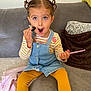 child, girl, pigtails, surprised, couch, yellow_leggings, striped_shirt, denim_dress, pink_comb, indoor, blanket, home, expression, playful, seated, curly_hair, furniture, cute, person, portrait