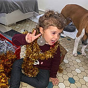 Naëlya participe au concours pour gagner de l'argent avec cette photo : toddler, child, smiling, peace_sign, gold_tinsel, christmas_ornament, curly_hair, dog, pet, indoors, floor_tiles, maroon_sweater, dark_pants, sock, holiday_decor, playful, happy, festive, home, curious_dog