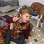 toddler, child, smiling, peace_sign, gold_tinsel, christmas_ornament, curly_hair, dog, pet, indoors, floor_tiles, maroon_sweater, dark_pants, sock, holiday_decor, playful, happy, festive, home, curious_dog