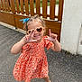 child, girl, sunglasses, heart_shaped, orange_dress, floral_pattern, outdoor, pavement, fence, playful, smiling, sandals, curly_hair, hair_accessory, posing, happy, summer, fashion, cute, portrait