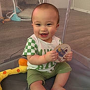 Kai is registered to the contest to win money with this photo: baby, child, smiling, playpen, toy, green_clothing, sitting, happy, infant, mesh_net, indoor, card, cute, toddler, shorts, shirt, person, skin, face, hand