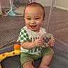 baby, child, smiling, playpen, toy, green_clothing, sitting, happy, infant, mesh_net, indoor, card, cute, toddler, shorts, shirt, person, skin, face, hand