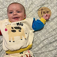 Weston is registered to the contest to win money with this photo: baby, infant, smiling, clothing, sweatshirt, animal_print, cow, blanket, pattern, wooden_sign, hand, face, cute, lying_down, child, happy, indoors, young, portrait, two_months